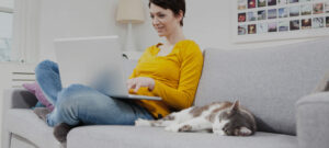 Frau sitzt auf Sofa mit Notebook und Katze liegt daneben