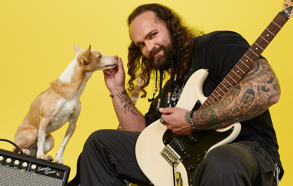 Ein rockiger Musiker ist mit seinem hellbraunen Hund beisammen. Im Hintergrund sieht man Verstärker und Gitarre.