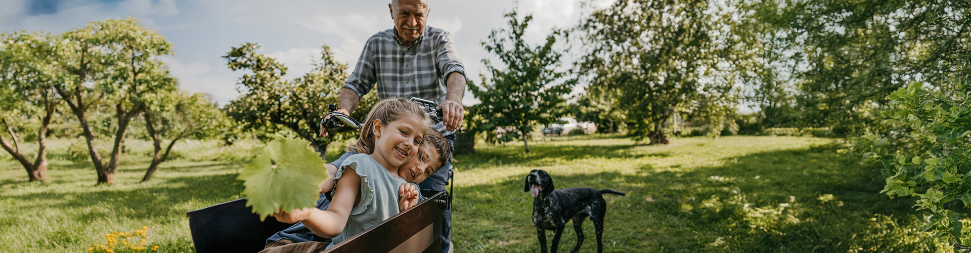 Zwei Kinder sitzen in einem Lastenrad, ein älterer Mann fährt dieses. Rund herum ist eine grüne Wiese mit einigen Bäumen und einem Hund.