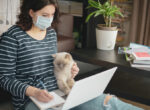 Eine Frau sitzt mit Maske an Ihrem Laptop. Ihre Katze sitzt auf ihrem Schoß. Vorsicht ist gefragt, da auch Katzen an Corona erkranken können.