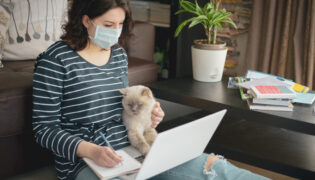 Eine Frau sitzt mit Maske an Ihrem Laptop. Ihre Katze sitzt auf ihrem Schoß. Vorsicht ist gefragt, da auch Katzen an Corona erkranken können.