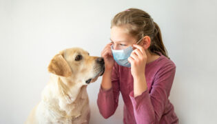 Ein Mädchen mit OP-Maske schaut ihrem hellen Hund in die Augen. Wie verändert sich durch das Coronavirus der Umgang mit dem vierbeinigen Begleiter?