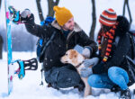 Mann und Frau sind mit ihrem Hund im Winterurlaub. Sie sitzen mit Snowboards und ihrem Hund im Schnee.