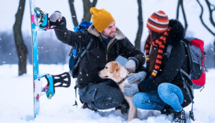 Mann und Frau sind mit ihrem Hund im Winterurlaub. Sie sitzen mit Snowboards und ihrem Hund im Schnee.