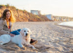 Frau sitzt mit Hund am Strand vor Dünen, sie besuchen gerne Hundestrände.