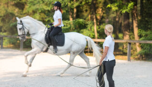 Reitlehrerin unterrichtet Reitschülerin auf weißem Pferd an der Longe