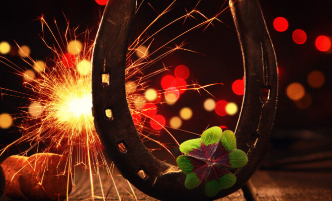 Hufeisen mit Sektkorken und einem vierblättrigen Kleeblatt sowie einer brennenden Wunderkerze im Hintergrund.