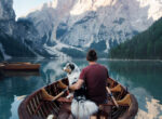 Mann sitzt mit Hund im Boot auf dunkelblauem See vor fantastischer, verschneiter Berglandschaft