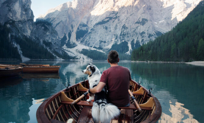 Mann sitzt mit Hund im Boot auf dunkelblauem See vor fantastischer, verschneiter Berglandschaft