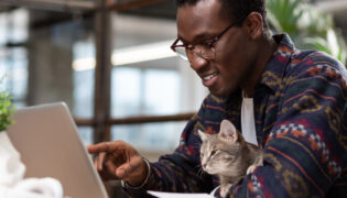 Mann schaut mit Katze auf einen Laptop. Wie viele D.N.A.-Übereinstimmungen haben Katzen und Menschen?