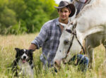 Mann mit Pferd und Reitbegleithund auf einer Wiese.