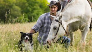 Mann mit Pferd und Reitbegleithund auf einer Wiese.