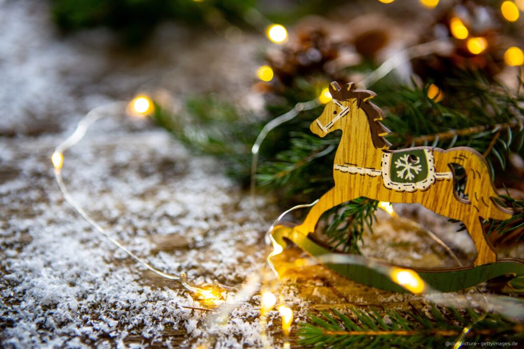 Ein braunes kleines Schaukelpferd steht auf einem mit Schnee bedecktem Stallboden vor einem Tannenzweig.