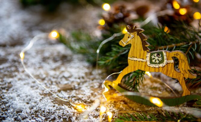 Ein braunes kleines Schaukelpferd steht auf einem mit Schnee bedecktem Stallboden vor einem Tannenzweig.