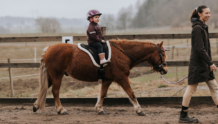 Reitlehrerin führt Kind auf Pferd über den Trainingsplatz.