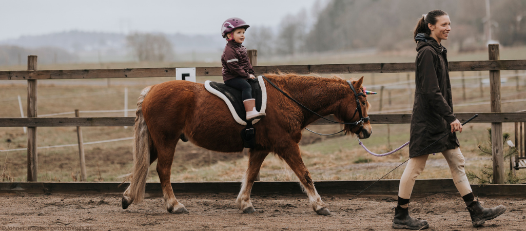 Reitlehrerin führt Kind auf Pferd über den Trainingsplatz.